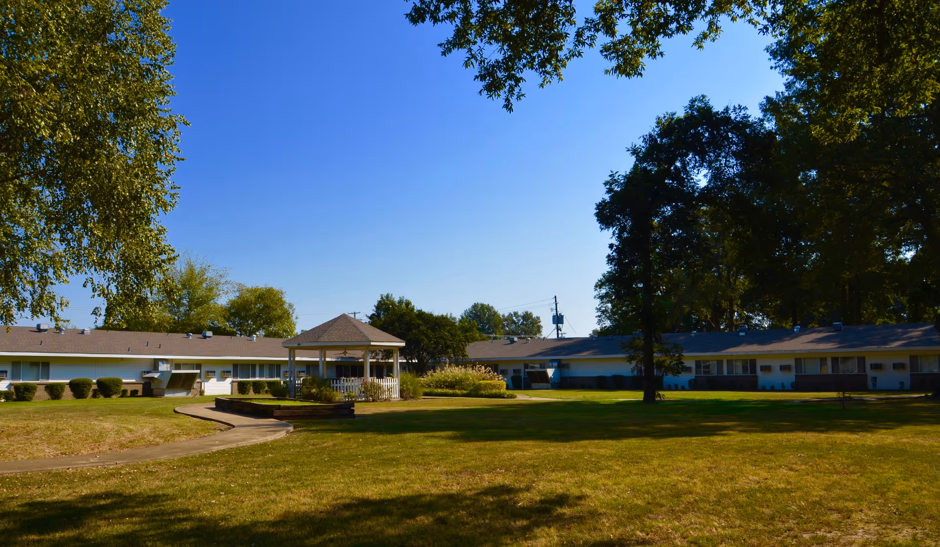 Outdoor view of Indianola Rehabilitation and Healthcare Center showing a large grassy lawn with a paved walkway leading to a white gazebo. The single-story building surrounds the lawn with trees providing shade and a clear blue sky overhead.