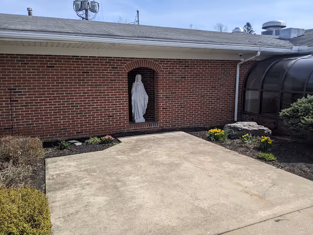 Outdoor view of a brick wall with a recessed niche containing a white statue. The area in front has a concrete patio surrounded by small flower beds with blooming yellow and pink flowers. There is a glass-covered walkway or sunroom attached to the right side of the building.