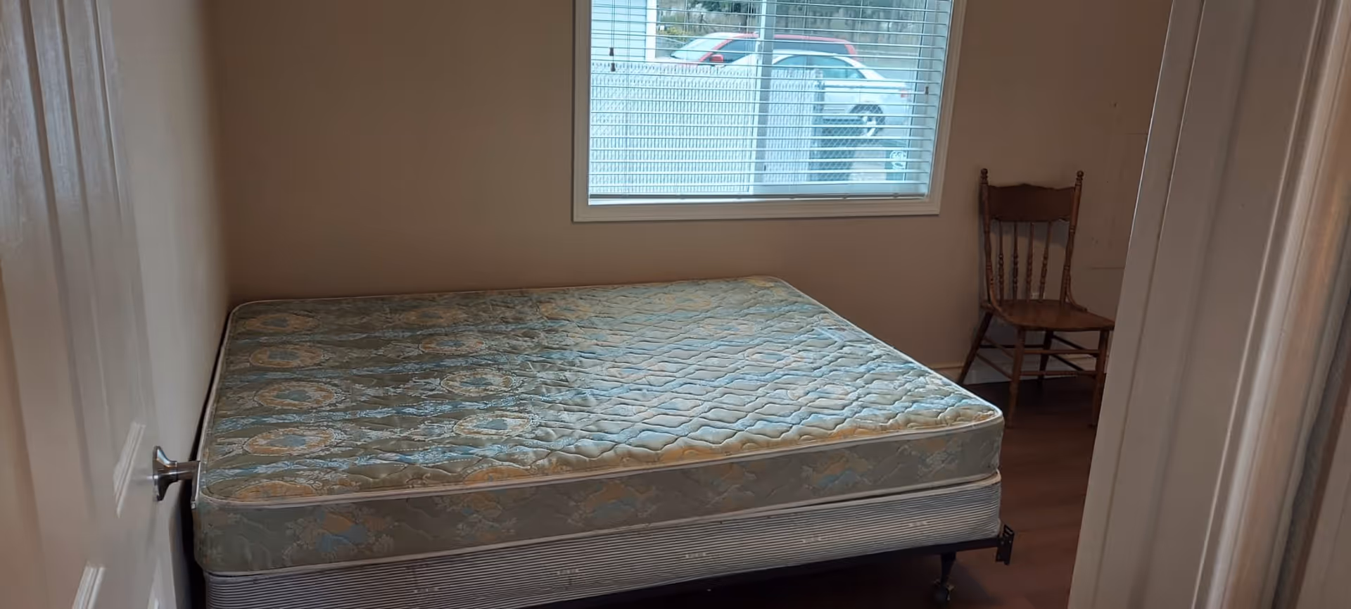 A small bedroom with a mattress on a bed frame positioned against the left wall. There is a window with blinds on the far wall, and a wooden chair is placed in the corner to the right of the window. The room has plain walls and wooden flooring.
