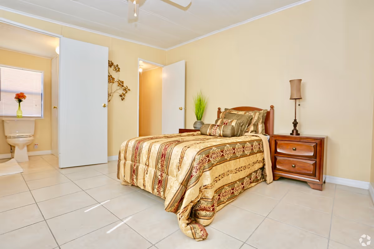 A bedroom with a single bed covered in a patterned gold and brown bedspread. Next to the bed is a wooden nightstand with two drawers and a lamp on top. The room has beige walls and tiled floors. Two open doors reveal a bathroom with a toilet and a window with blinds, and a hallway with warm lighting. A decorative wall piece is visible between the doors.