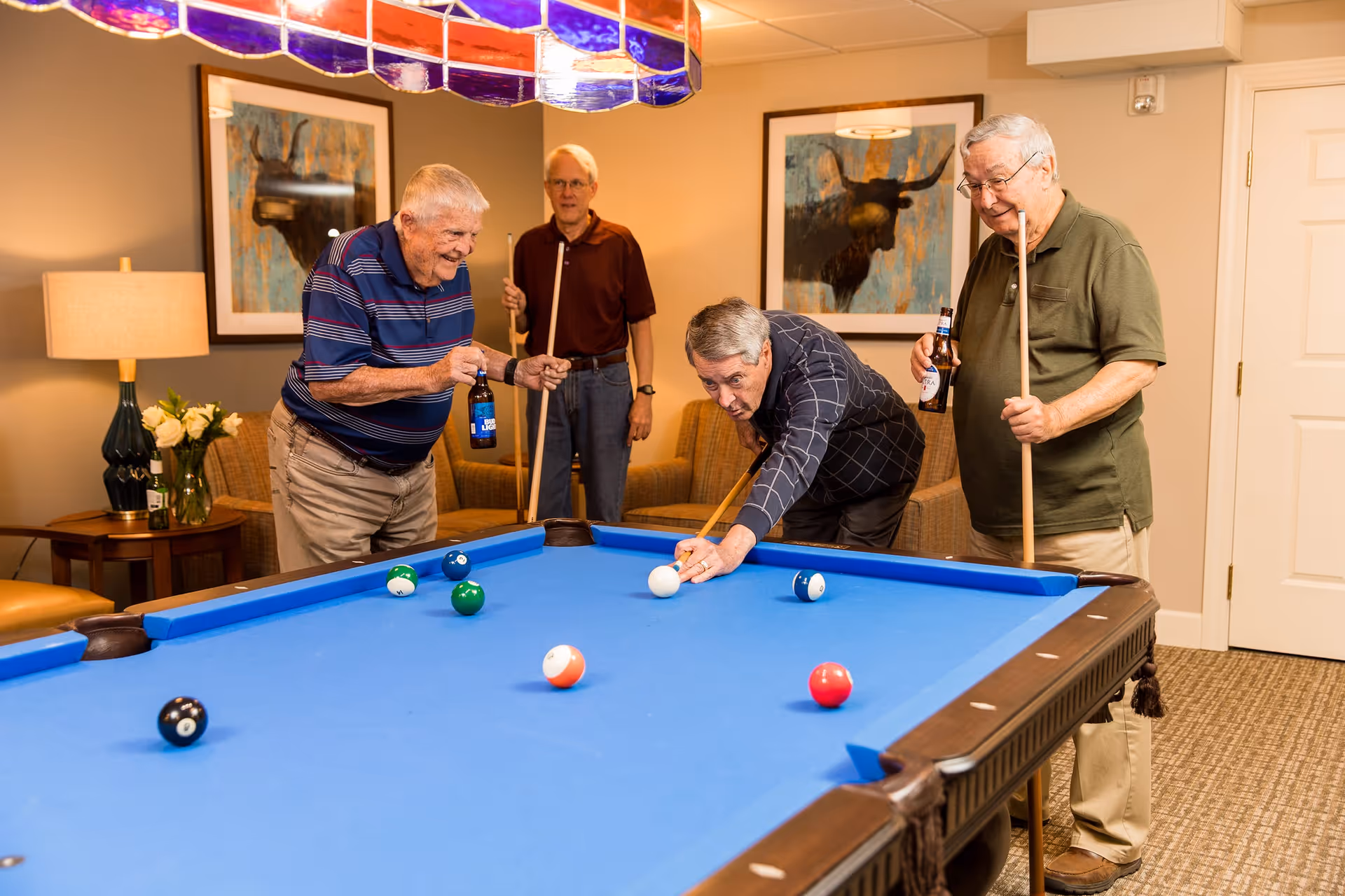 Four elderly men playing pool in a cozy room with beige walls, two framed paintings of bulls, and a stained glass light fixture above the pool table. Two men are holding pool cues and bottles of beer, while one man is leaning over the table aiming a shot with a cue stick. The room has comfortable seating and a side table with a lamp and flowers.