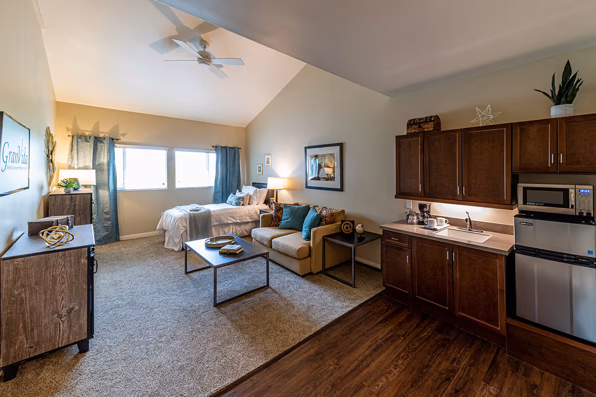 A spacious senior living studio apartment with a bed near three large windows covered with blue curtains. The room features a beige sofa with colorful pillows, a coffee table, a side table with a lamp, and a kitchenette with wooden cabinets, a sink, microwave, and mini refrigerator. The walls are light-colored, and the floor is a combination of carpet and wood. A ceiling fan is mounted on the vaulted ceiling.