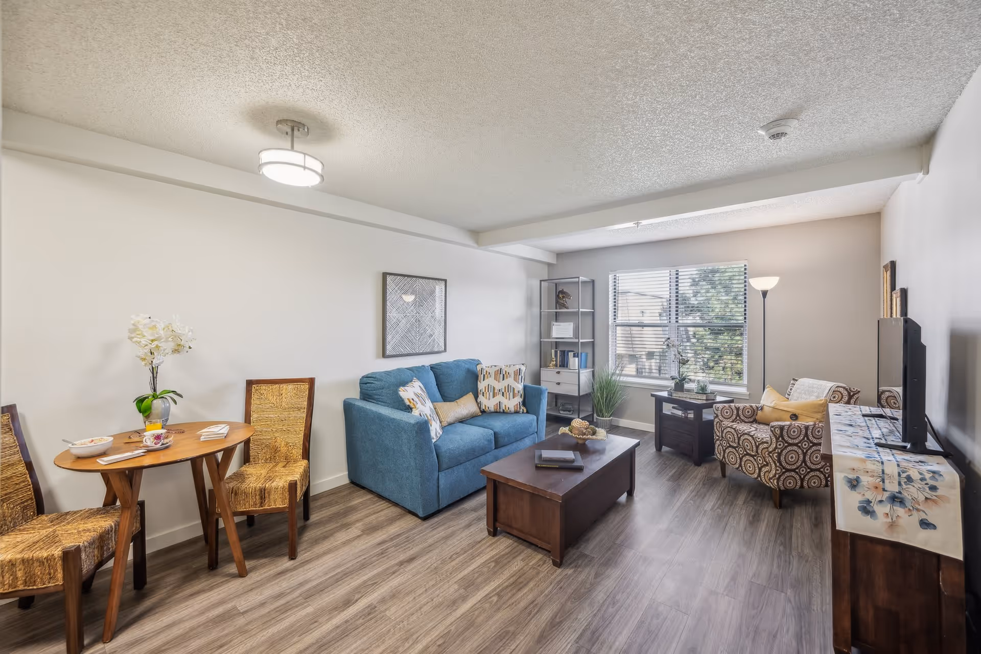 A bright living room with wood flooring featuring a blue loveseat with patterned cushions, a wooden coffee table, a patterned armchair, a small round wooden dining table with two wicker chairs, a tall floor lamp, a window with blinds, and a TV on a wooden stand with a floral runner.