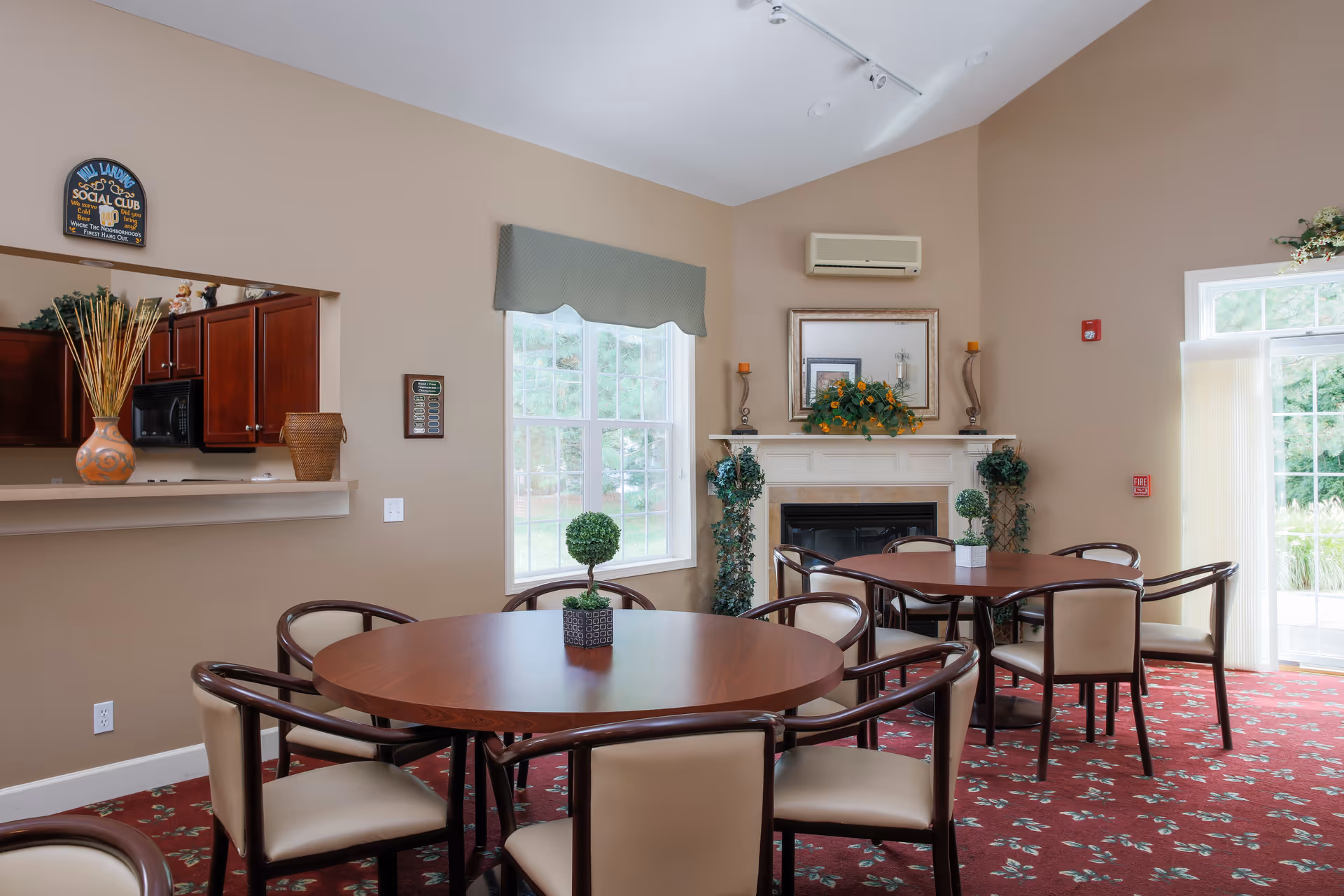A cozy common area with round wooden tables and cushioned chairs on a red floral carpet. There is a fireplace with decorative plants and a framed mirror above it. Large windows let in natural light, and a kitchen pass-through window is visible on the left side.