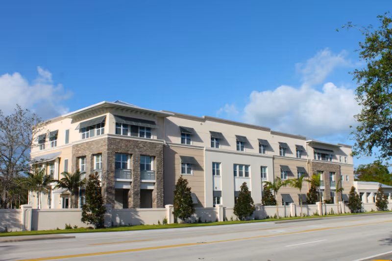 Three-story light-colored senior living building with stone accents facing a street under a blue sky.