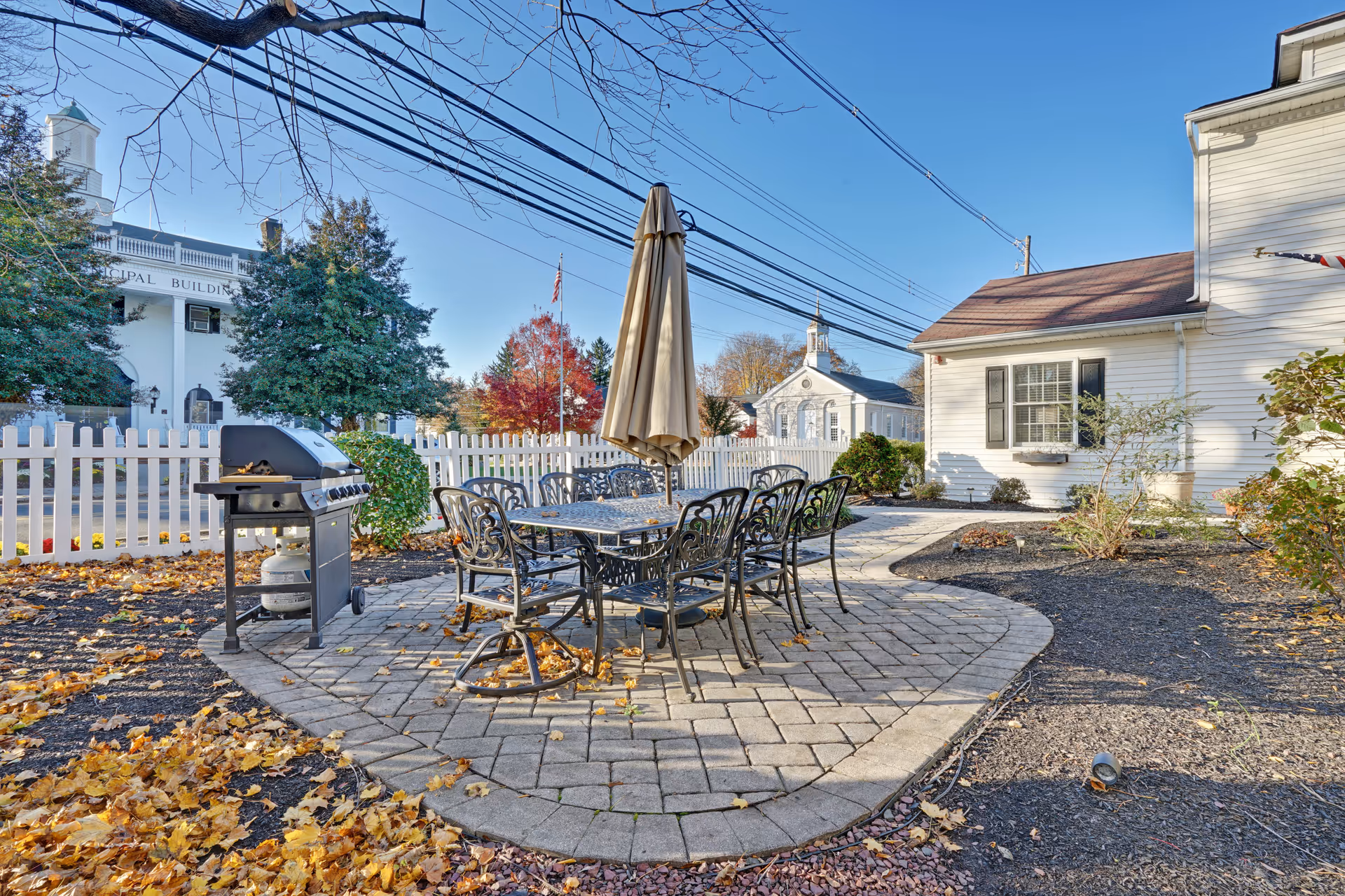 Outdoor patio area with a metal table and eight matching chairs arranged around it. A closed beige umbrella stands in the center of the table. To the left of the table is a gas grill. The patio is paved with stone tiles and surrounded by a garden area with fallen autumn leaves. In the background, there is a white picket fence, trees with fall foliage, and white buildings under a clear blue sky.