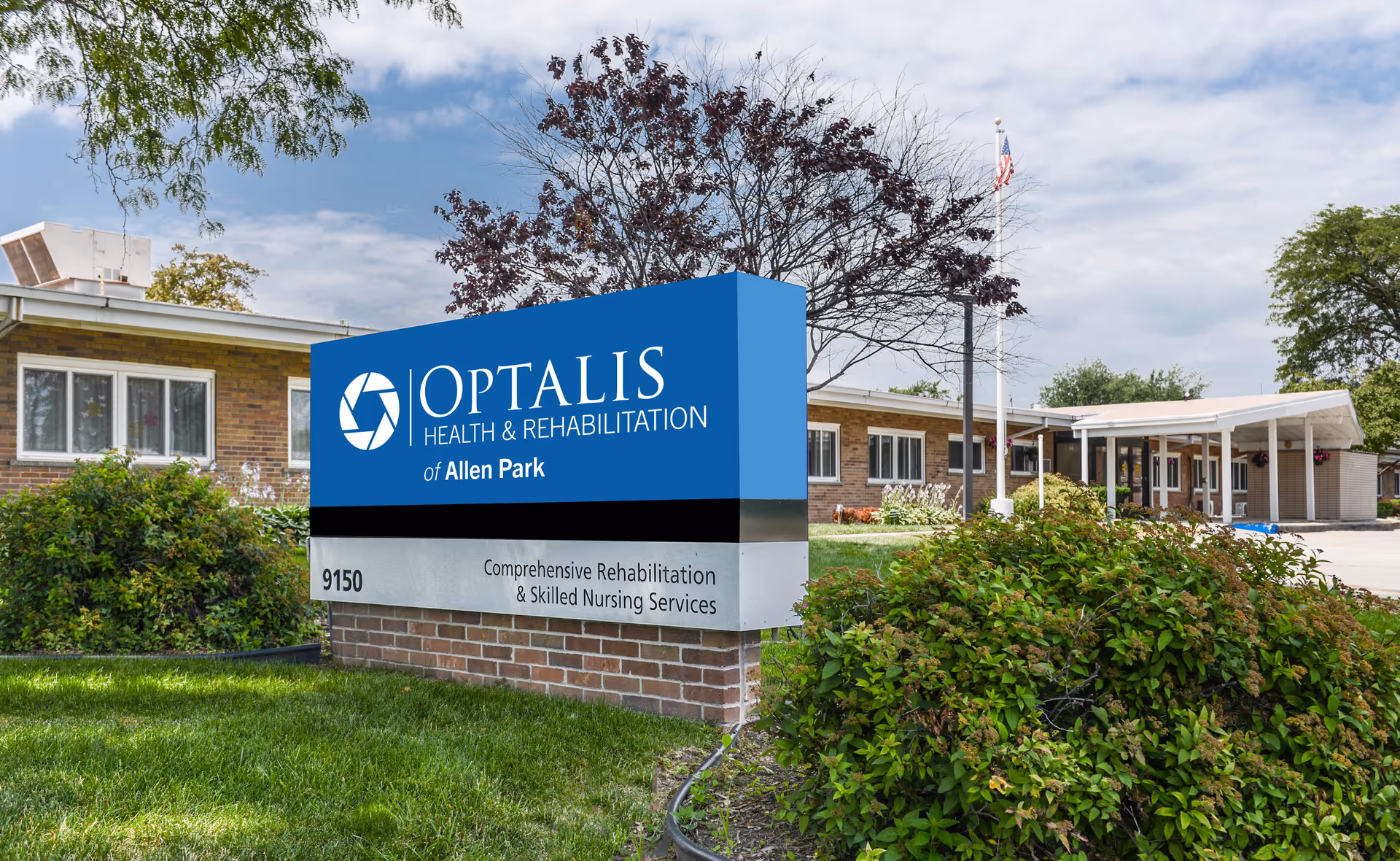 Exterior view of Optalis Health & Rehabilitation of Allen Park facility showing a large blue and gray sign with the facility name and services, surrounded by green bushes and grass, with the building and an American flag in the background under a partly cloudy sky.