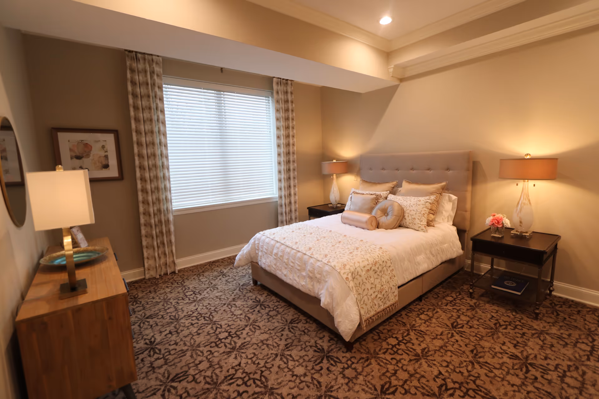 A cozy bedroom with a beige upholstered bed adorned with multiple decorative pillows and a patterned bed runner. The room features a large window with blinds and patterned curtains, a wooden dresser with a lamp and decorative items, and a nightstand with a lamp and a small flower arrangement. The carpet has a floral pattern, and the walls are painted a soft beige color.