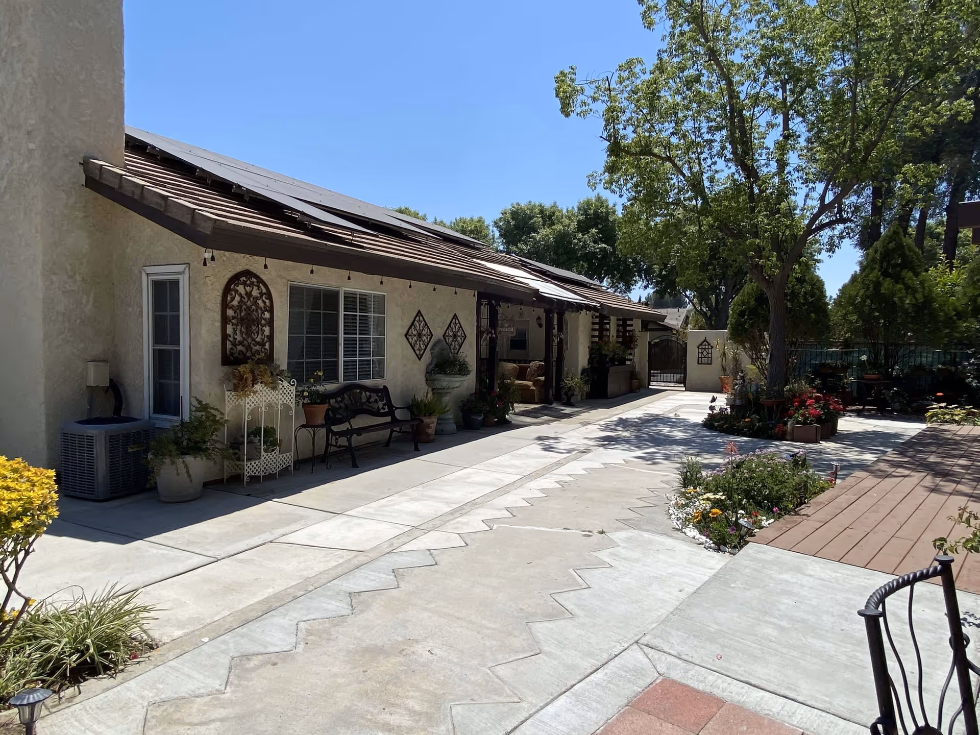 Outdoor courtyard area of Cypress Residence featuring a paved walkway with zigzag patterns, a bench, potted plants, decorative wall hangings, a tree providing shade, and a gated entrance in the background under a clear blue sky.