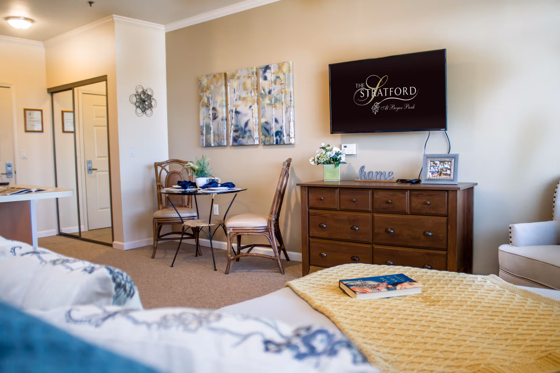 Sunlit furnished senior apartment room with a bed in the foreground, a small dining table and chairs, and a dresser with a wall-mounted TV displaying the Stratford logo.