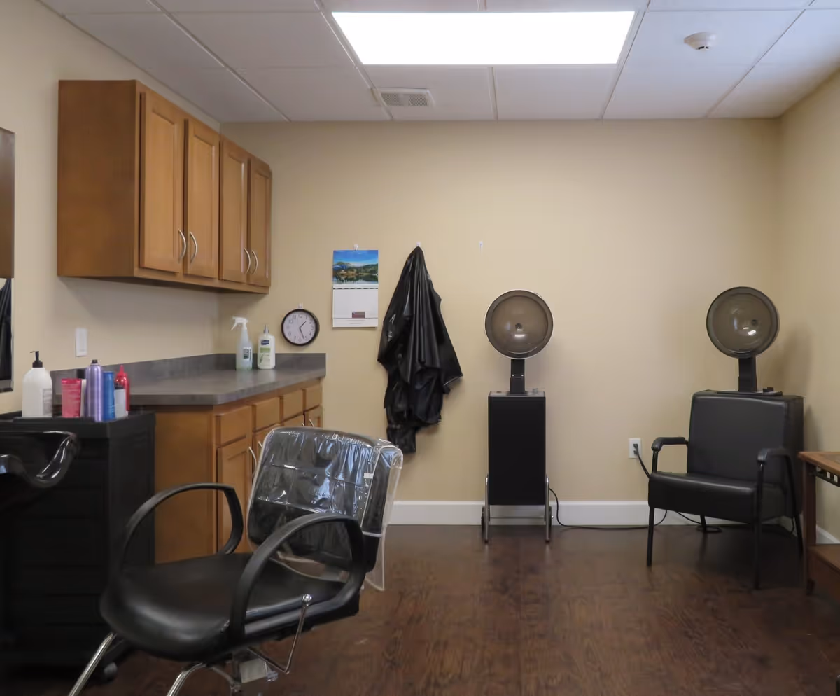 Interior of a hair salon area in a senior living facility with two black salon chairs, two hair dryers, wooden cabinets on the wall, various hair care products on a counter, a clock, and a calendar on a beige wall.
