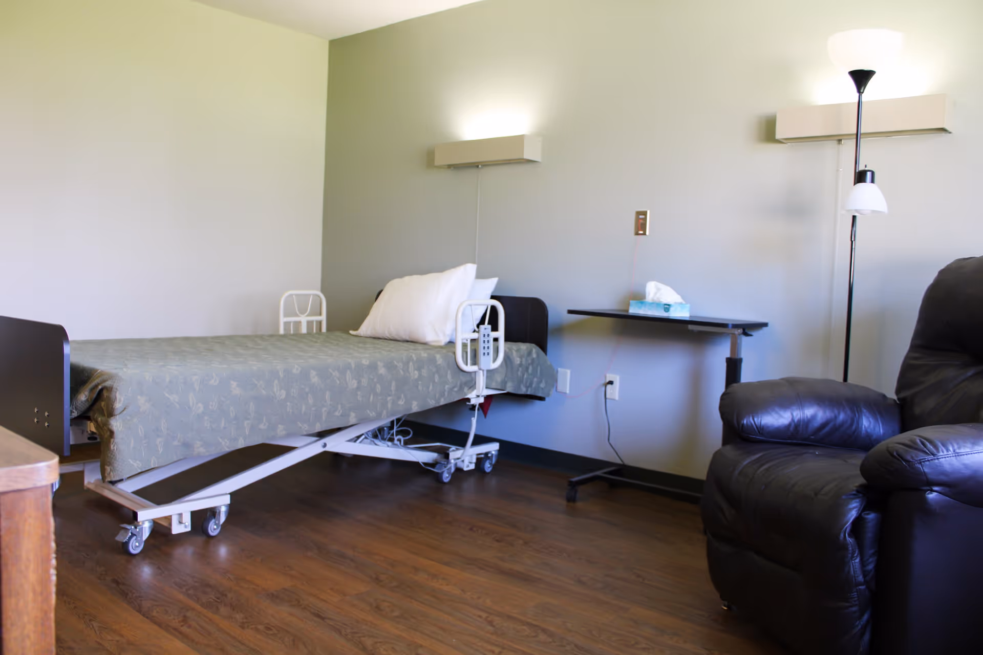 A simple senior care bedroom with a hospital-style bed, bedside table with tissues, floor lamp, and a leather recliner.