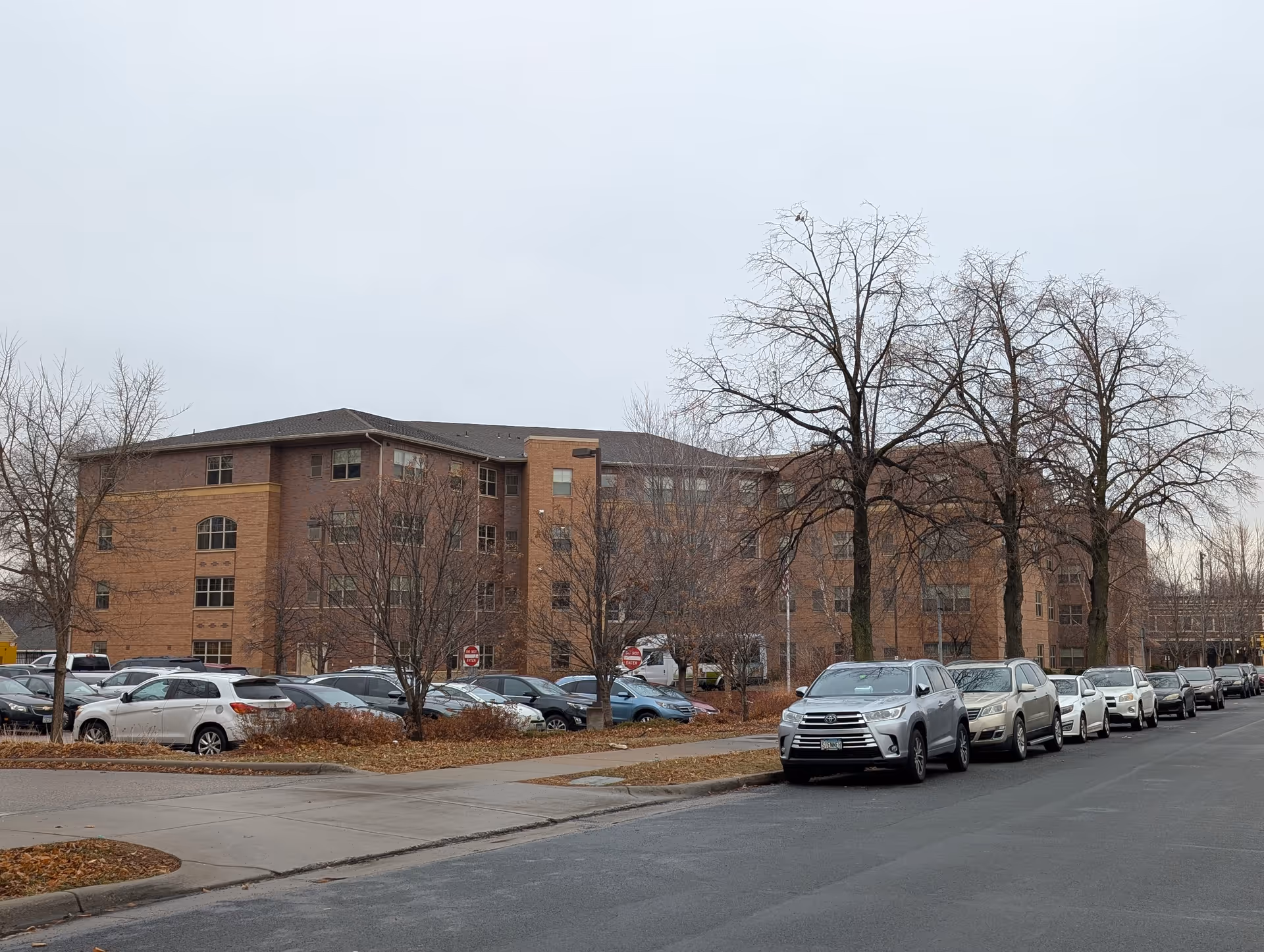 Brick four-story senior living building seen from the street with parked cars and leafless trees on an overcast day.