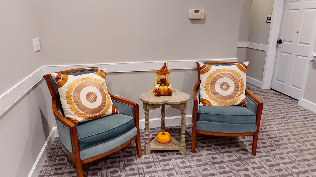 Two upholstered armchairs with orange patterned pillows flank a small round side table holding fall decorations and a pumpkin on a carpeted hallway floor.