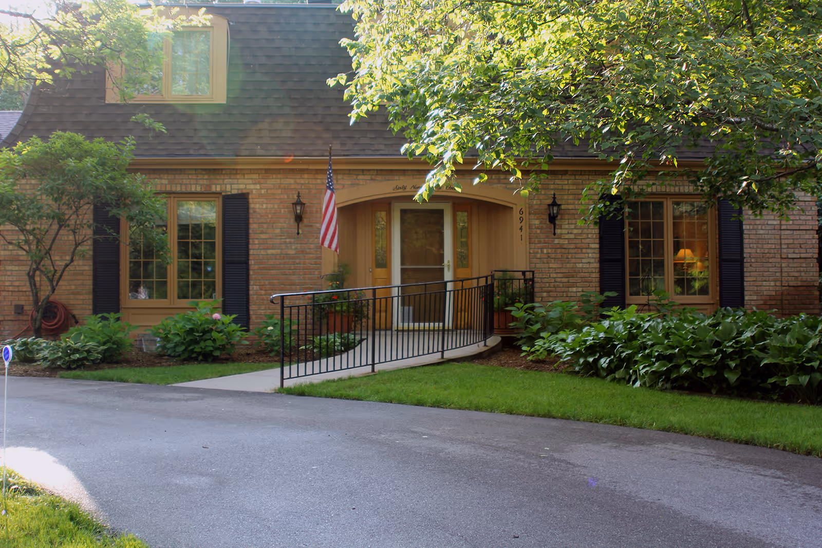 Exterior view of a brick building with a sloped roof, two large windows with black shutters, and a central entrance with a ramp and handrails. There is an American flag mounted near the door, surrounded by green shrubs and trees.