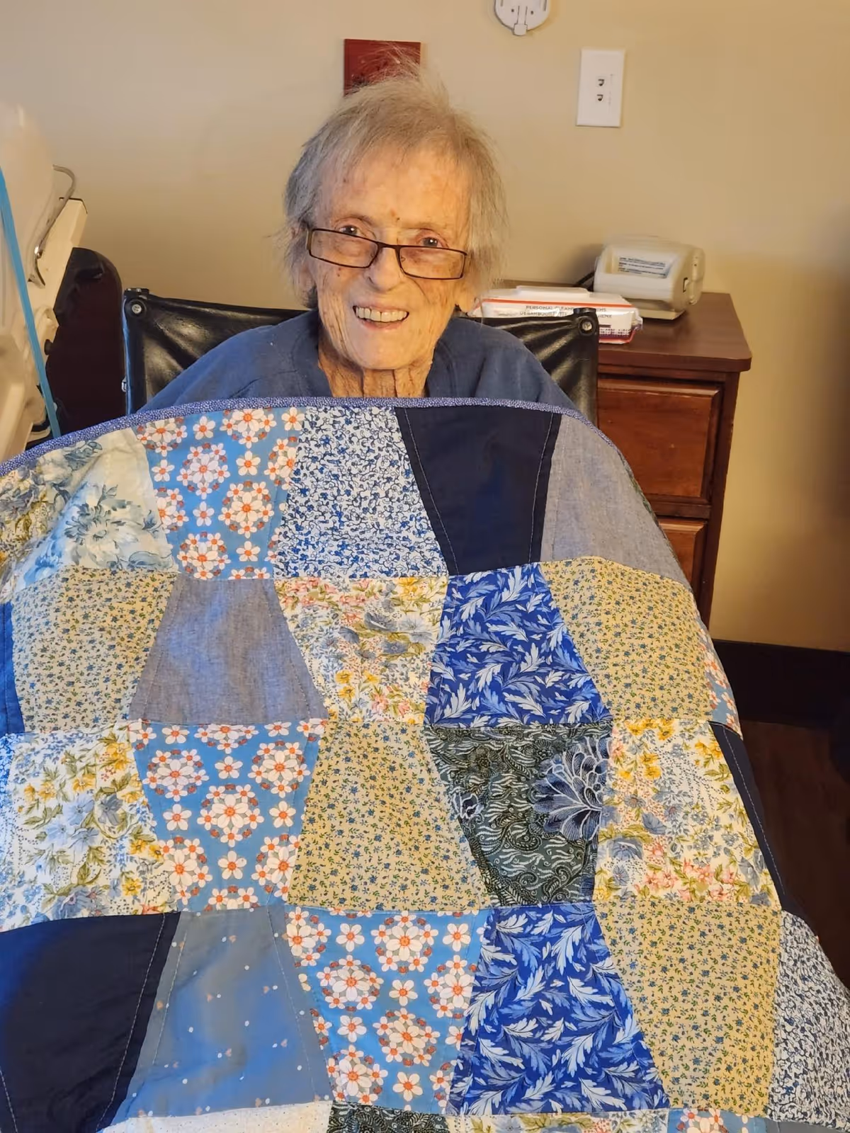 An elderly woman with glasses is sitting in a wheelchair, smiling and covered with a colorful patchwork quilt. Behind her is a wooden nightstand with a telephone and some items on it, and a beige wall with electrical outlets.