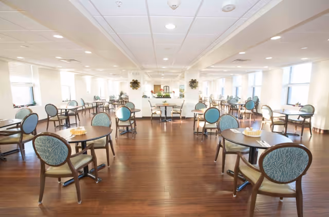 A spacious, well-lit dining room with multiple round tables and chairs arranged neatly. The room has large windows allowing natural light to fill the space, wooden flooring, and a clean, modern ceiling with recessed lighting. Each table has a small basket and utensils placed on it.