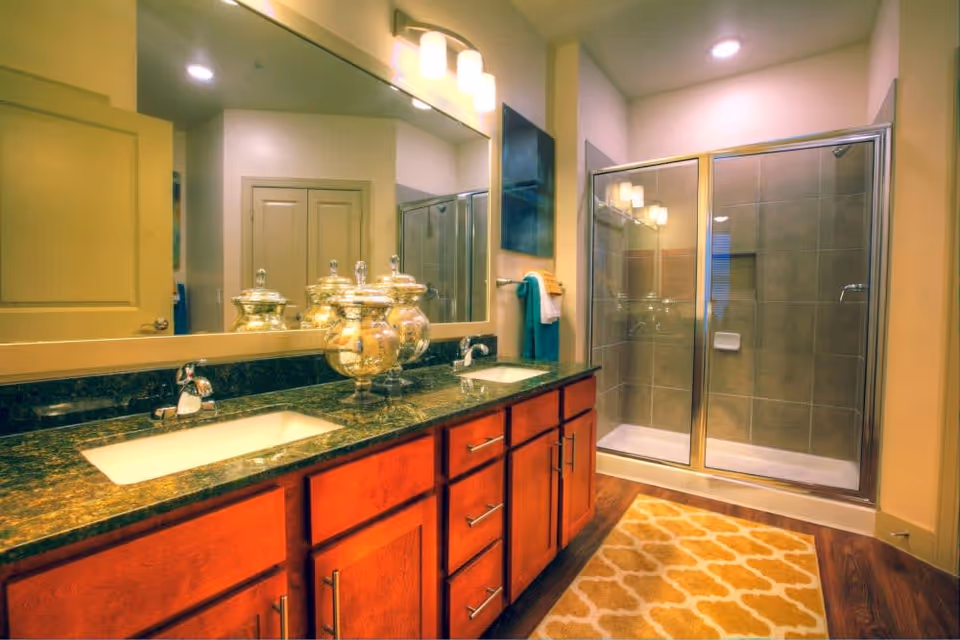 A bathroom featuring a double sink vanity with dark green marble countertops and wooden cabinets. Above the sinks is a large mirror with three light fixtures. To the right is a glass-enclosed shower with beige tiles. A patterned yellow and white rug is placed on the wooden floor in front of the shower.