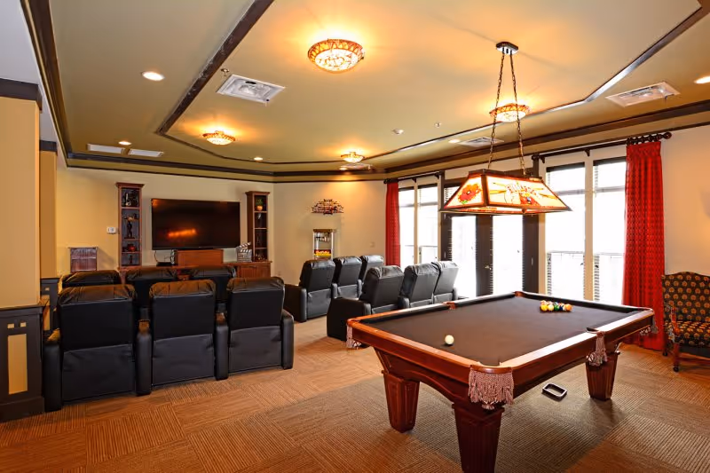 A cozy entertainment room with a pool table in the foreground and two rows of black leather recliners facing a large flat-screen TV mounted on the wall. The room has warm lighting with ceiling fixtures and a hanging lamp above the pool table. Red curtains frame the windows on the right side, and there is a patterned armchair in the corner.