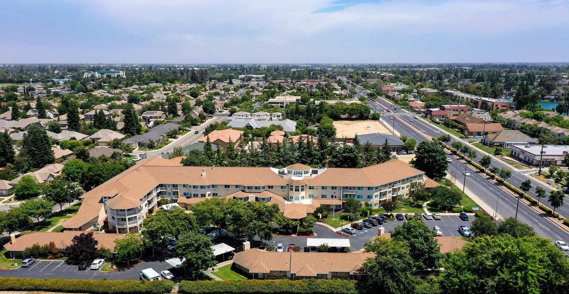 Aerial exterior view of a large curved senior living facility building with parking lots, trees, and surrounding neighborhood.