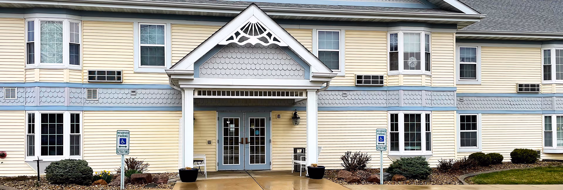 Front entrance of a two-story pale yellow building with a covered portico, double glass doors, and reserved handicapped parking signs.