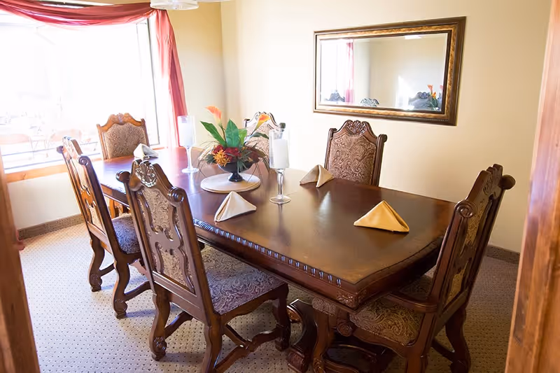 A dining room with a wooden table set for four, featuring ornate carved chairs with patterned upholstery. The table has folded napkins, two candle holders, and a floral centerpiece. A large window with red curtains lets in natural light, and a framed mirror hangs on the beige wall.