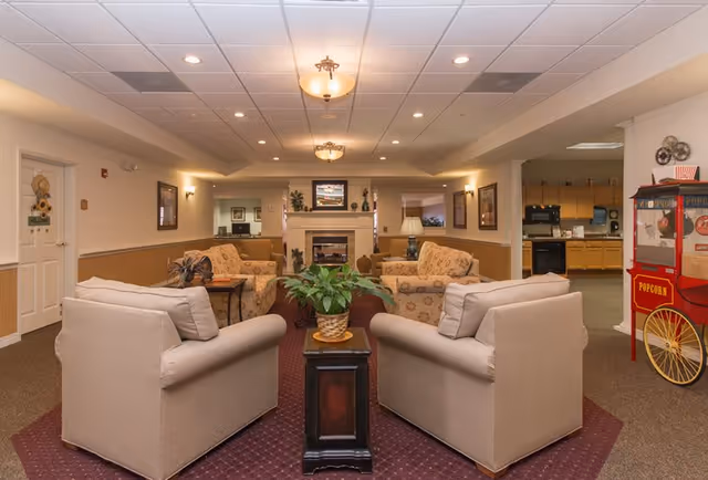 A cozy senior living facility common area with beige armchairs and floral sofas arranged around a central fireplace. A small wooden table with a potted plant is placed between two armchairs in the foreground. The room has warm lighting with ceiling fixtures and wall sconces. To the right, there is a vintage-style popcorn machine and a kitchen area visible in the background.