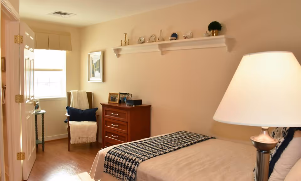 A cozy bedroom with a bed covered in a beige blanket and a black and white patterned throw. A bedside lamp with a white shade is on the right. Across from the bed is a wooden dresser with framed photos on top. A wooden chair with a white throw blanket and a dark blue pillow is positioned near a window with a beige valance. Above the dresser, a white wall shelf holds decorative items including small plants and figurines. The room has light-colored walls and wooden flooring.
