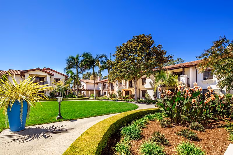 Sunny outdoor courtyard area at Smith Health Care Center at Covenant Living at the Samarkand featuring a well-maintained green lawn, palm trees, flowering plants, and a paved walkway surrounded by shrubs and landscaping. Two-story buildings with balconies and tiled roofs are visible in the background under a clear blue sky.