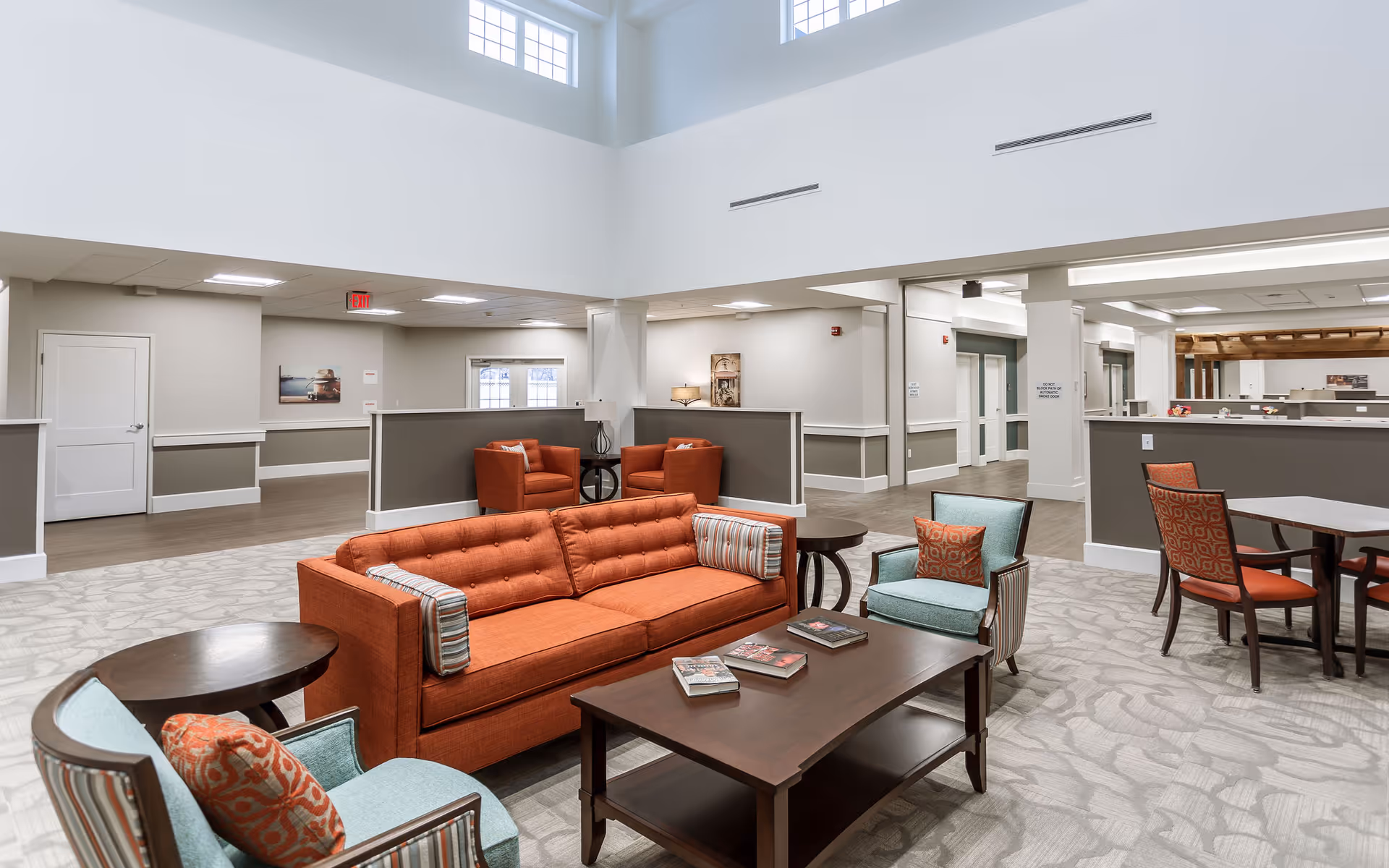 A spacious and well-lit senior living facility common area with high ceilings and large windows. The room features an orange sofa with striped pillows, two teal armchairs with patterned cushions, a dark wooden coffee table with magazines, and additional seating areas with orange chairs and tables. The walls are painted in neutral tones with white trim, and the floor is covered with a light patterned carpet.
