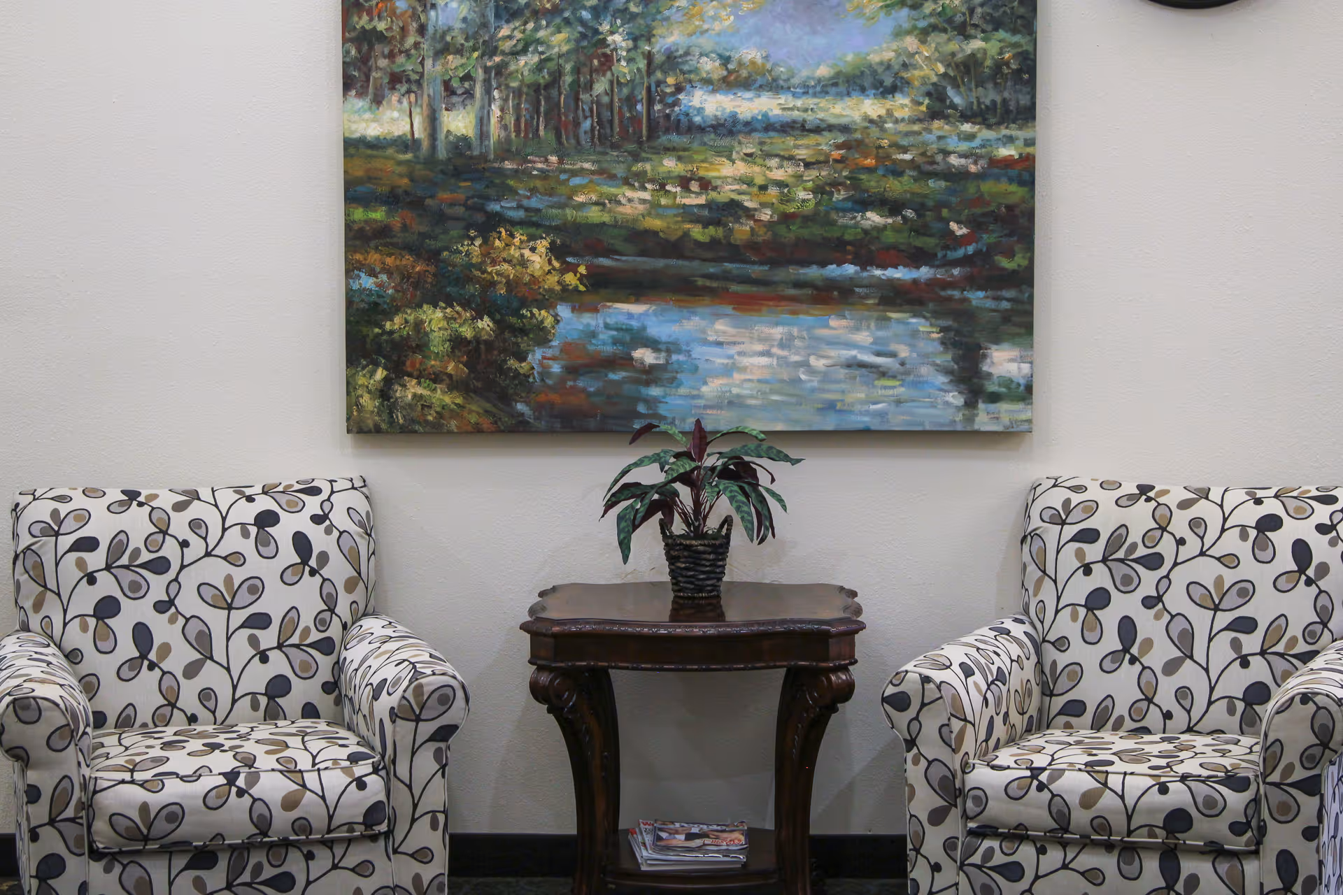 Two patterned armchairs with a floral vine design are placed on either side of a dark wooden side table holding a potted plant. Above the table hangs a large painting depicting a serene landscape with trees and a pond.