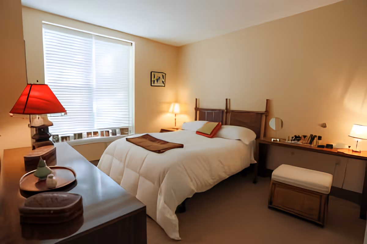 A cozy bedroom with a large bed covered in a white comforter and a brown throw blanket. The room features a wooden headboard, a bedside table with a lamp, a window with white blinds, a wooden dresser with decorative items, and a small cushioned bench in front of a vanity desk with a mirror and another lamp.