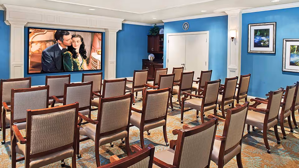 A small theater room with rows of wooden chairs facing a large screen mounted on a blue wall. The screen displays a scene from a classic movie featuring a man and a woman. The room has framed artwork on the walls and a carpeted floor with a patterned design.