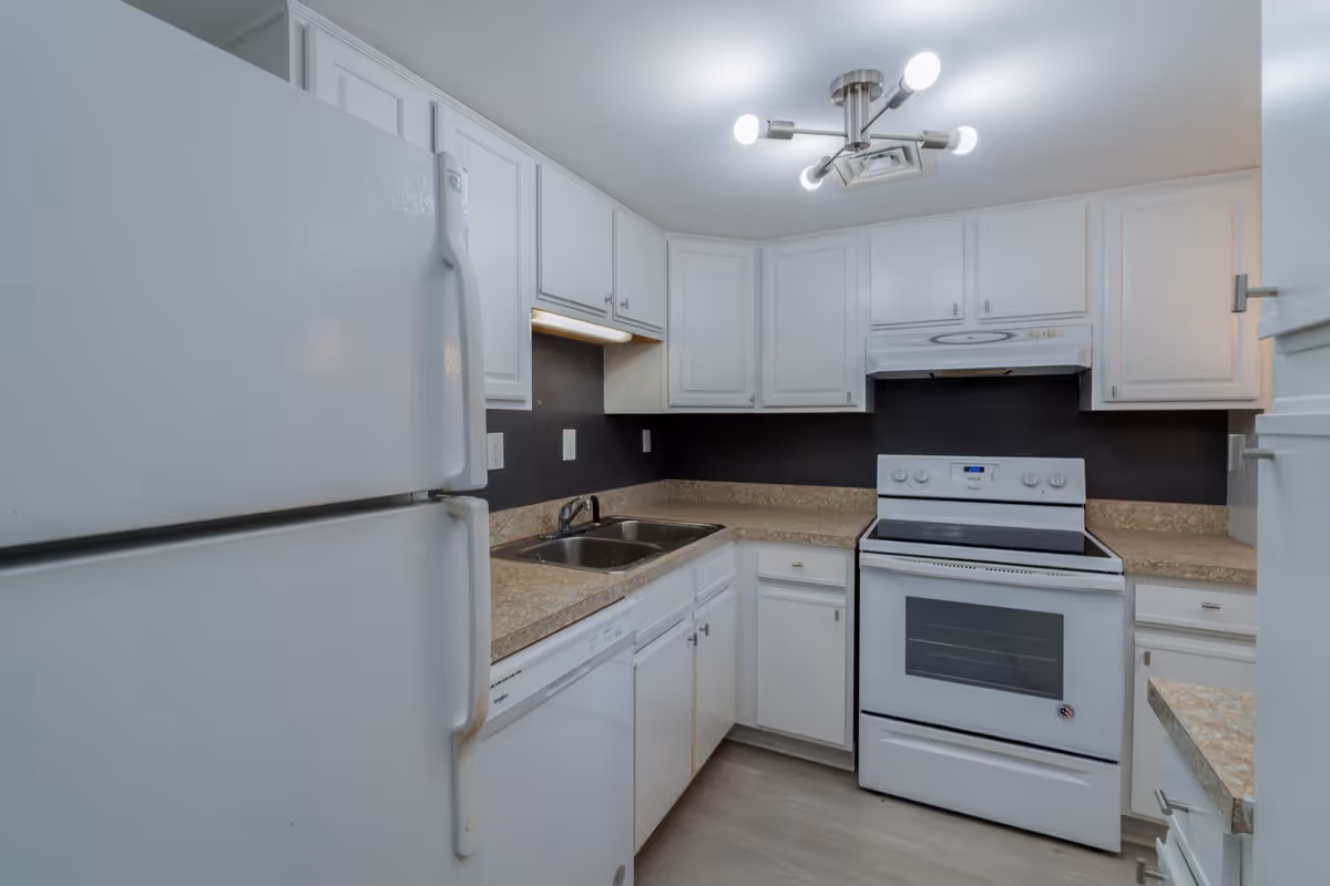 A compact kitchen with white cabinets, a white refrigerator, a white electric stove with an oven, a double sink, a dishwasher, and beige countertops. The kitchen has a modern ceiling light fixture and a black backsplash.