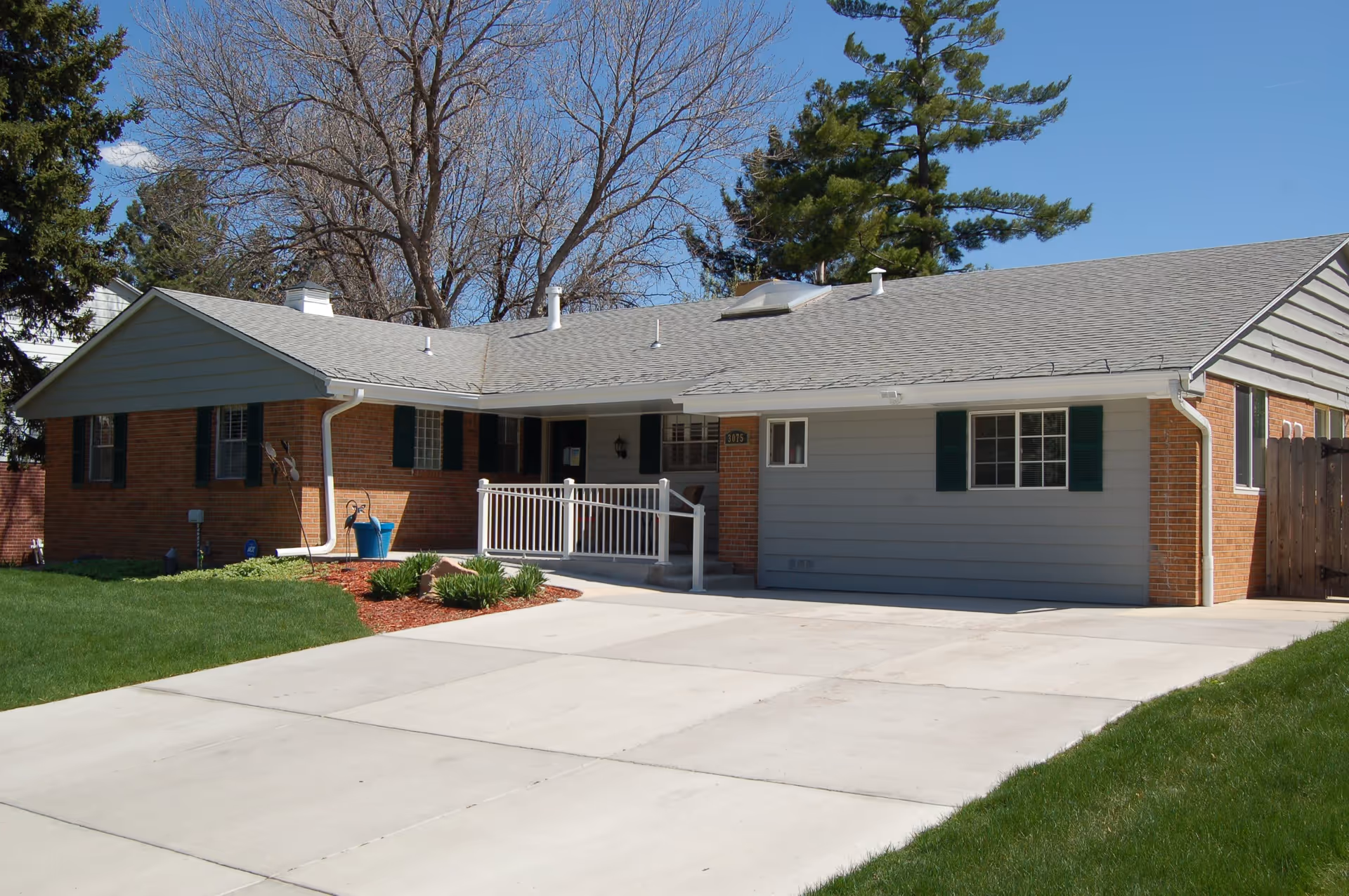 Single-story residential building with a combination of brick and gray siding exterior, green shutters on windows, a concrete driveway, a small front porch with a white railing, and a well-maintained lawn and landscaping under a clear blue sky.