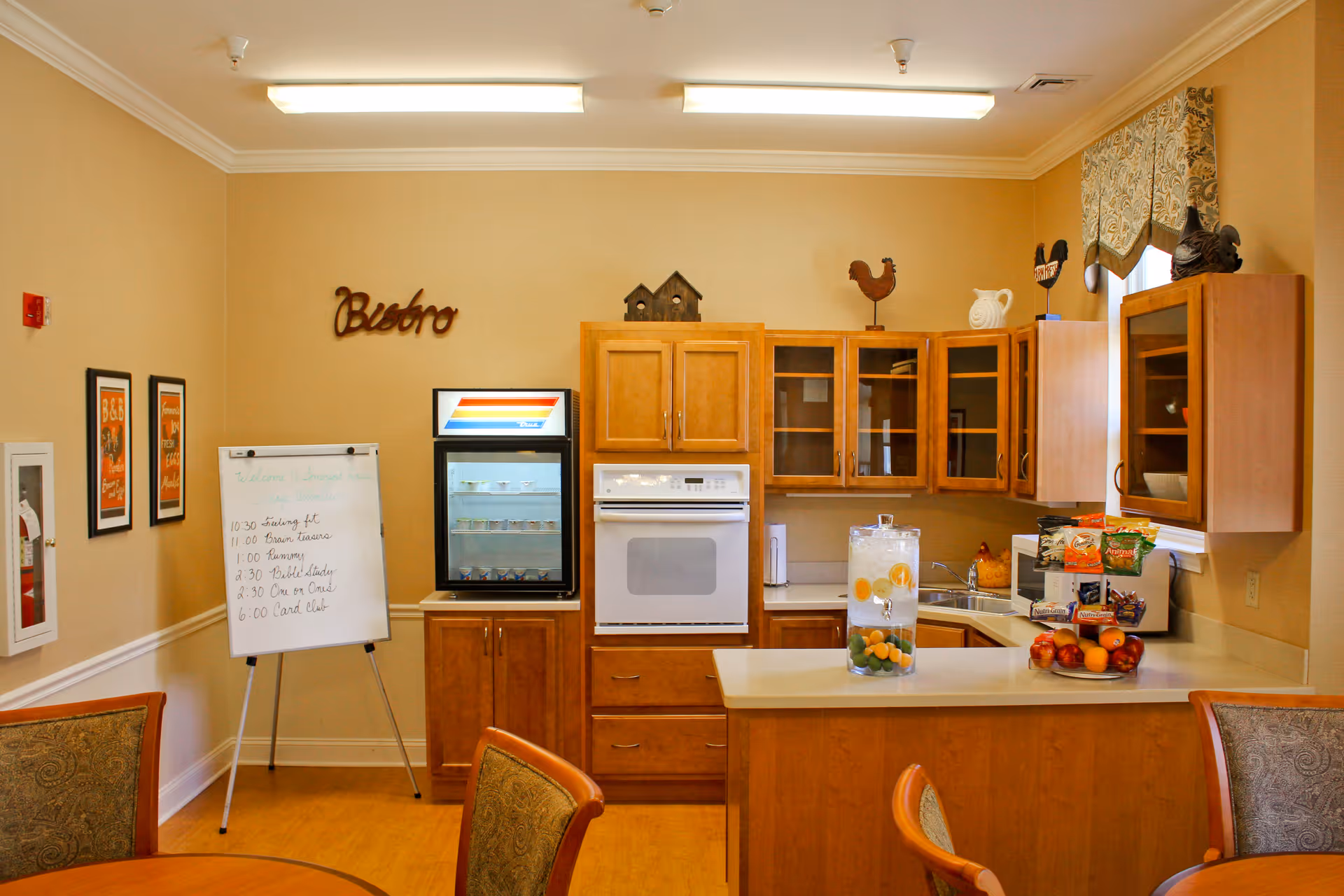 A cozy kitchen area in a senior living facility with wooden cabinets, a built-in oven, a small refrigerator, and a countertop with a glass dispenser filled with water and fruit. There is a whiteboard with a schedule, decorative items on top of the cabinets, and a round table with chairs in the foreground.