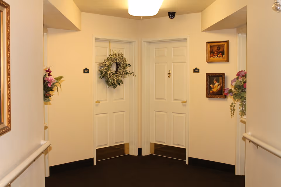 Interior hallway of a senior living facility with two white doors labeled 301 and 302. Door 301 has a decorative green wreath, and door 302 has two framed paintings on the wall beside it. There are floral arrangements on both sides of the hallway and a ceiling light fixture above.