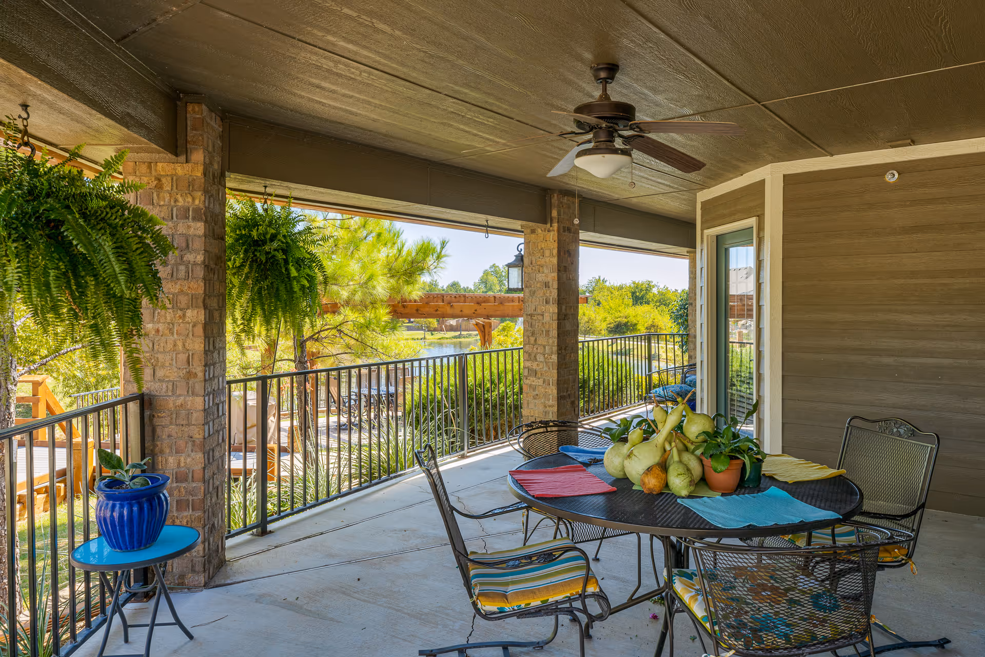 Covered outdoor patio area with a round metal table and four chairs with colorful cushions. The table has a centerpiece of gourds and a small potted plant. Two hanging ferns are suspended from the ceiling. The patio overlooks a garden with trees and a body of water in the background.