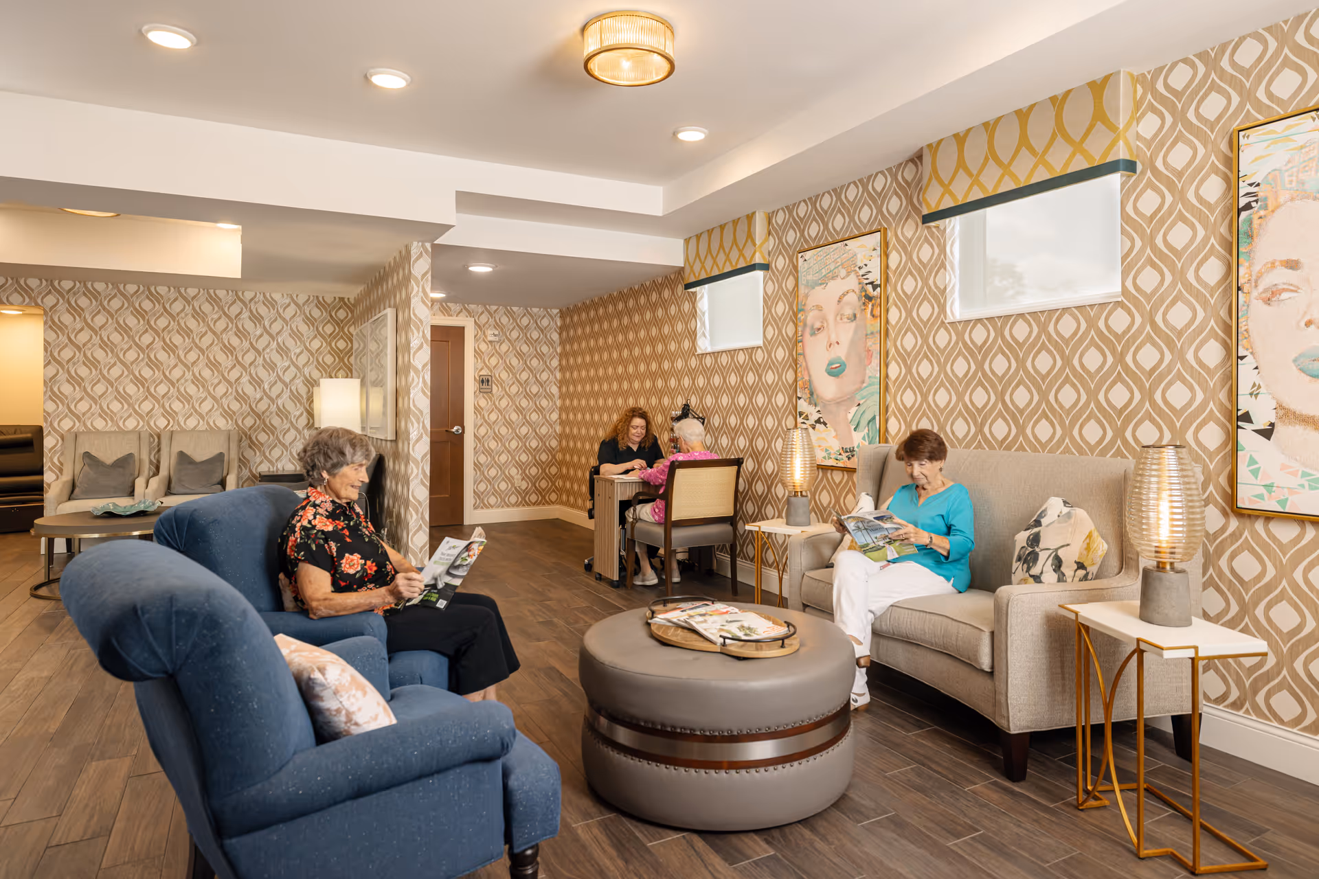 A cozy senior living facility lounge area with two elderly women sitting on comfortable chairs and sofas, reading magazines. The room features patterned wallpaper, modern lighting fixtures, framed artwork on the walls, and a round ottoman in the center. In the background, two more women are seated at a small table engaged in conversation.
