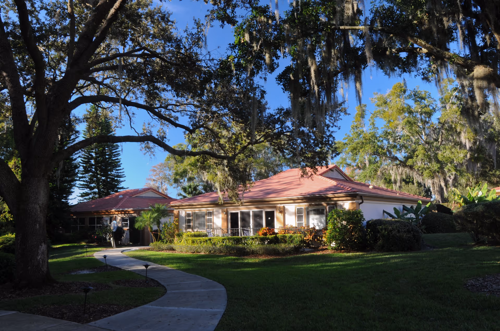 Single-story red-roofed building set among large oak trees with a curved walkway leading to the entrance.