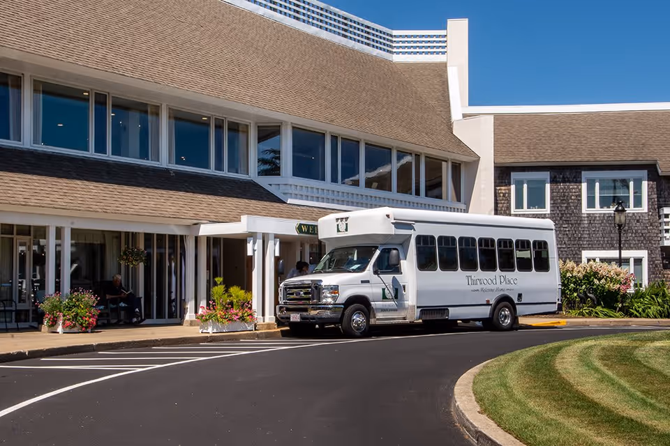 Exterior view of Thirwood Place showing a white shuttle bus parked in front of the building entrance. The building has large windows, a sloped roof, and landscaped greenery with flowers near the entrance. The sky is clear and blue.