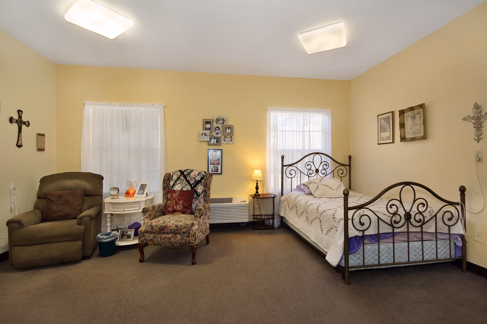 A cozy bedroom with a metal-framed bed covered with a white quilt, two armchairs including a floral patterned one and a brown recliner, a small white side table with decorative items, framed photos on the wall, and two windows with sheer white curtains letting in natural light.