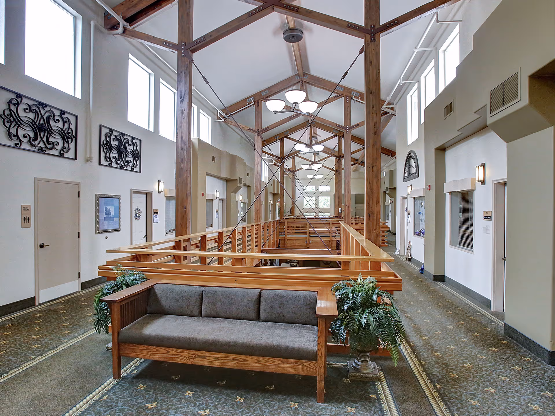 Interior view of a senior living facility hallway with a high vaulted ceiling supported by wooden beams and metal cables. There is a wooden bench with gray cushions and two potted plants on either side in the foreground. The hallway has multiple doors, windows near the ceiling, decorative wall art, and carpeted floors.