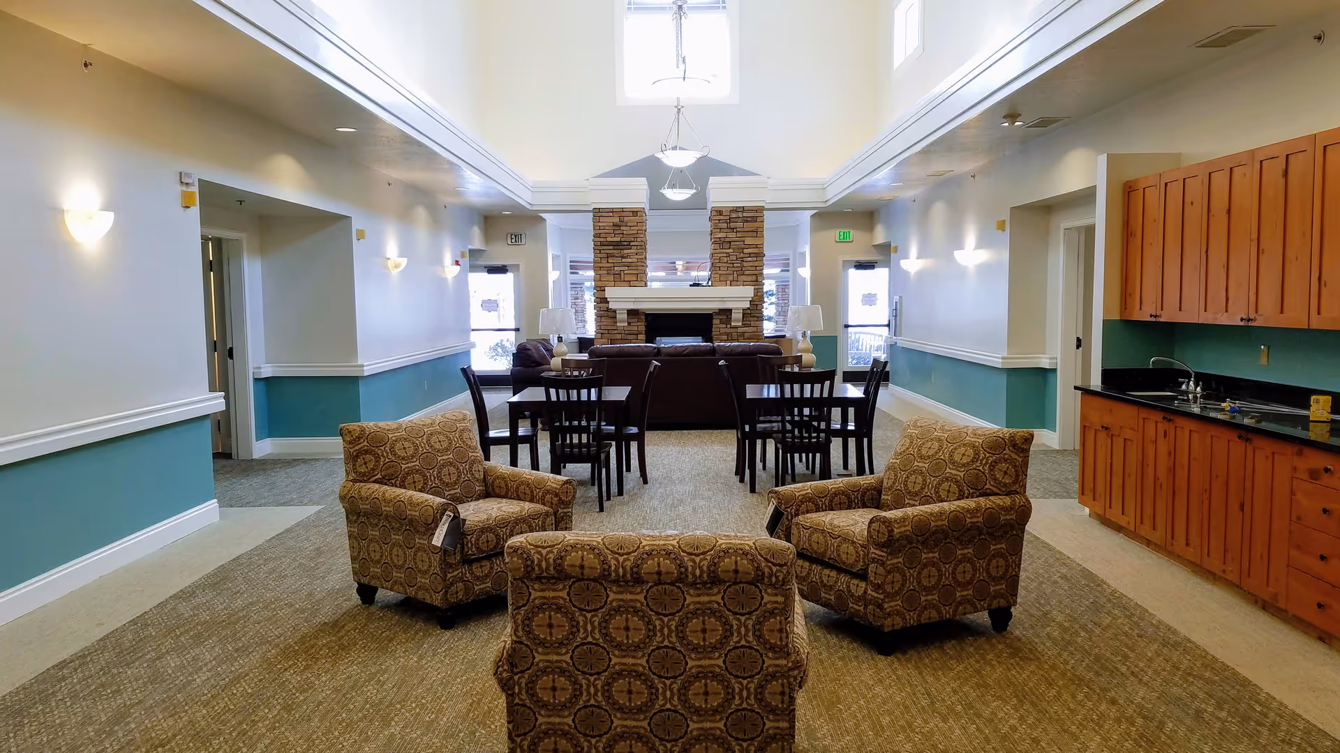 Spacious senior living common room featuring patterned armchairs, dining tables, a stone fireplace and a kitchenette.