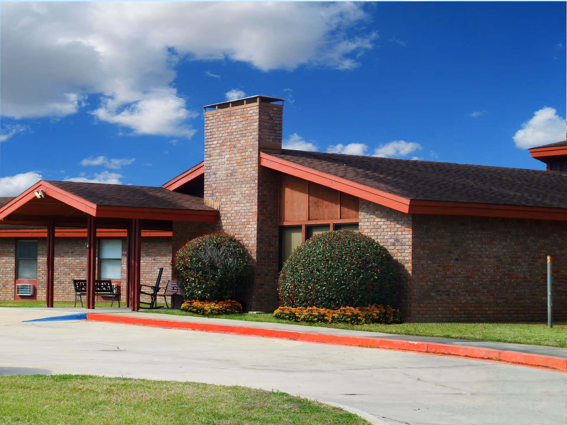 Exterior view of a single-story brick building with a chimney, red trim, and a covered entrance. There are two large bushes with flowers at the base in front of the building, a concrete driveway with a red curb, and a bright blue sky with scattered clouds.