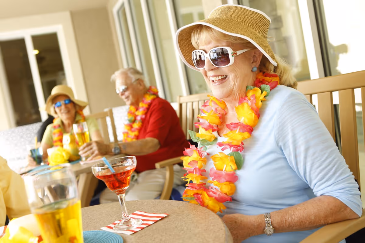 A group of elderly people sitting outdoors at a table, wearing colorful Hawaiian leis and sun hats, enjoying drinks and smiling.