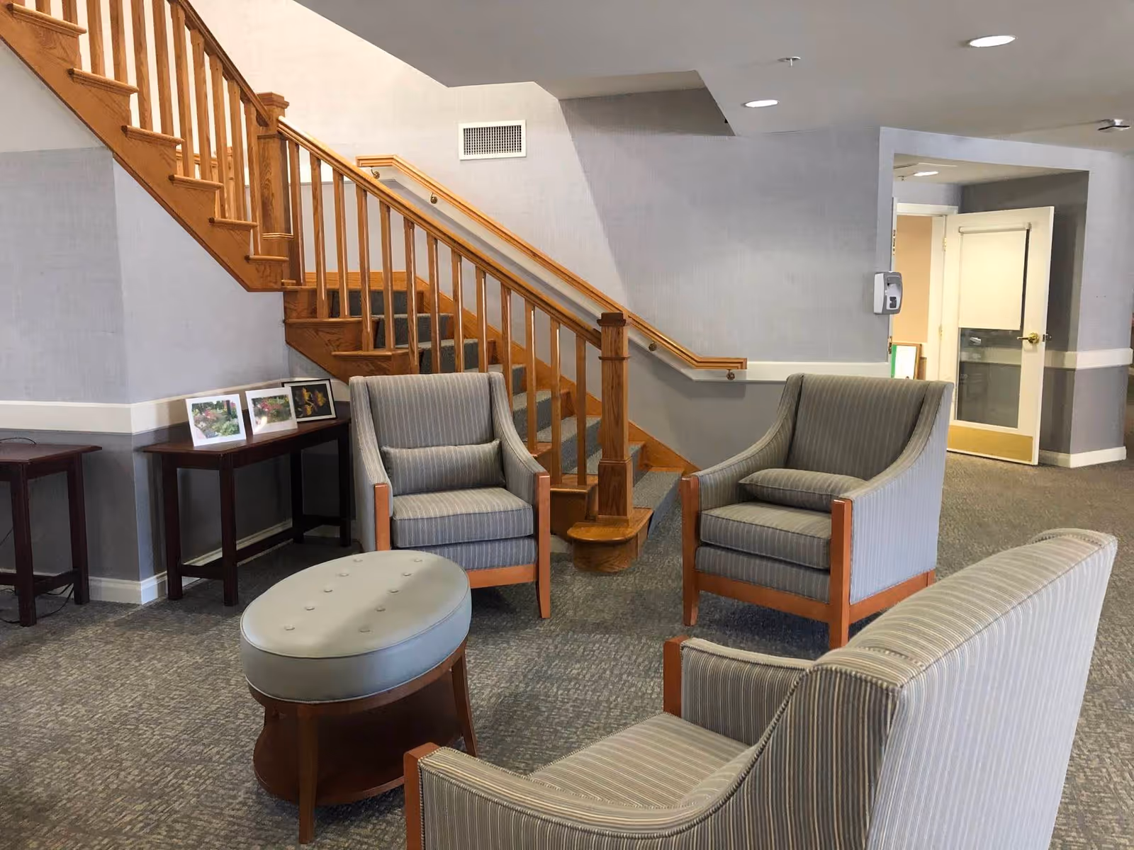 A lounge-style common area with striped armchairs, a round ottoman, a wooden staircase and side tables.
