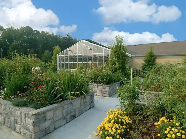 Raised stone garden beds and flowering plants leading to a glass greenhouse attached to a building under a blue sky.