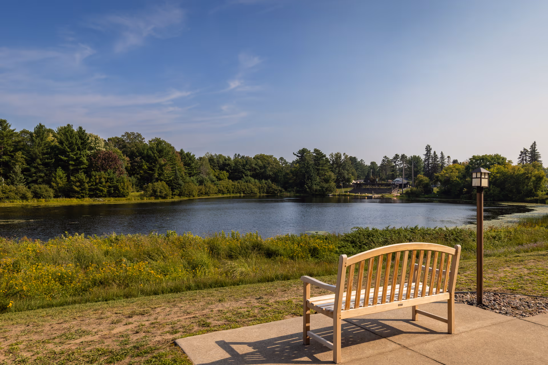 A wooden bench on a concrete patio facing a calm lake surrounded by green trees and vegetation under a clear blue sky.