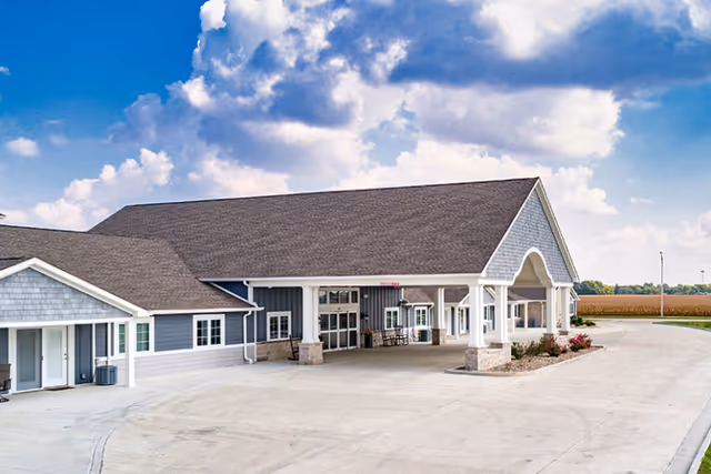 Exterior view of a single-story assisted living facility building with a large covered entrance supported by white columns. The building has gray siding with white trim and a dark shingled roof. The surrounding area includes a paved driveway and some landscaping with flowers. The sky is partly cloudy with blue patches.